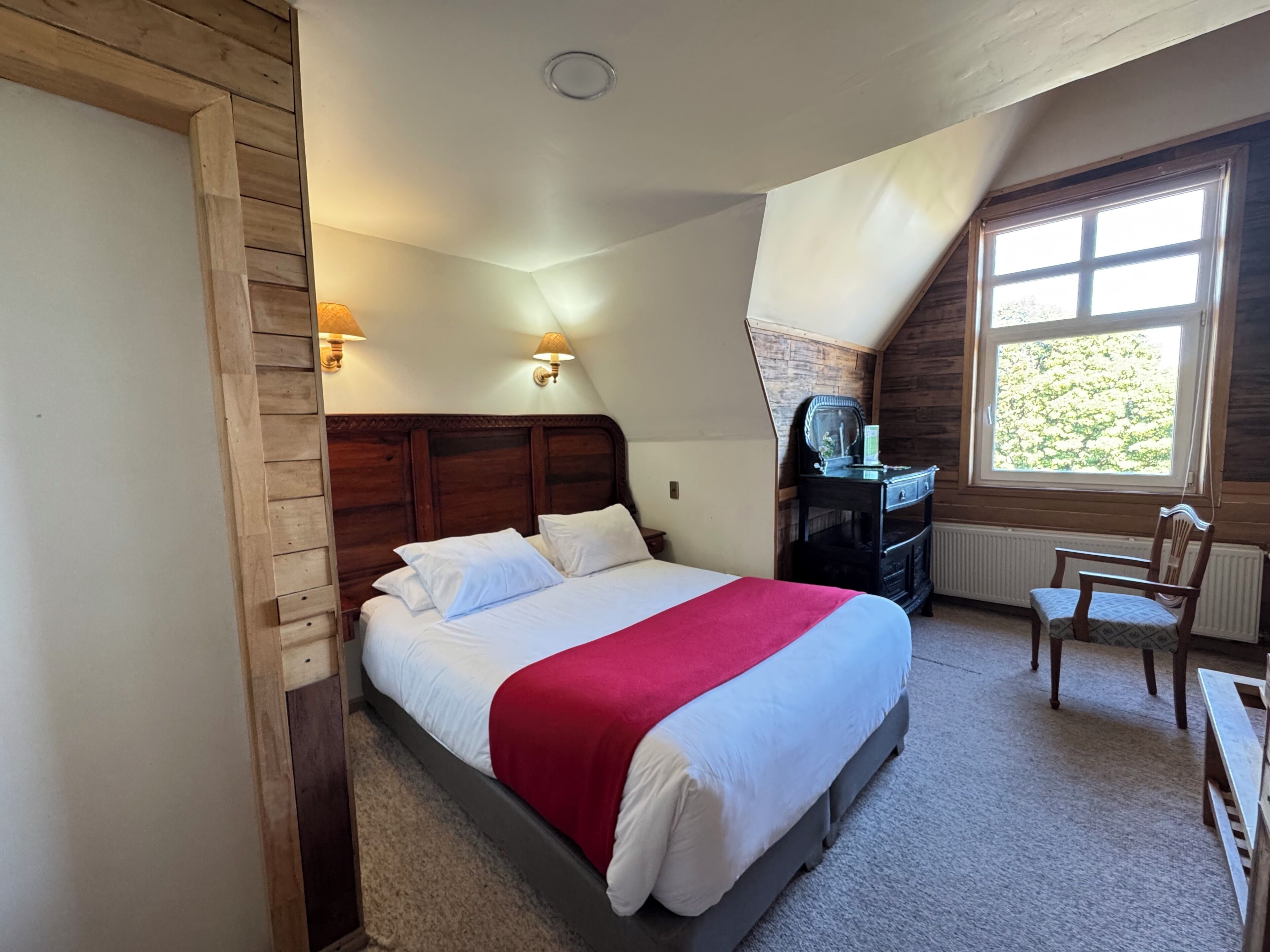 Un dormitorio con cabecera de madera, cama con ropa de cama blanca y una colcha roja, mesita de noche, cómoda, una silla y una ventana con vistas a zonas verdes.