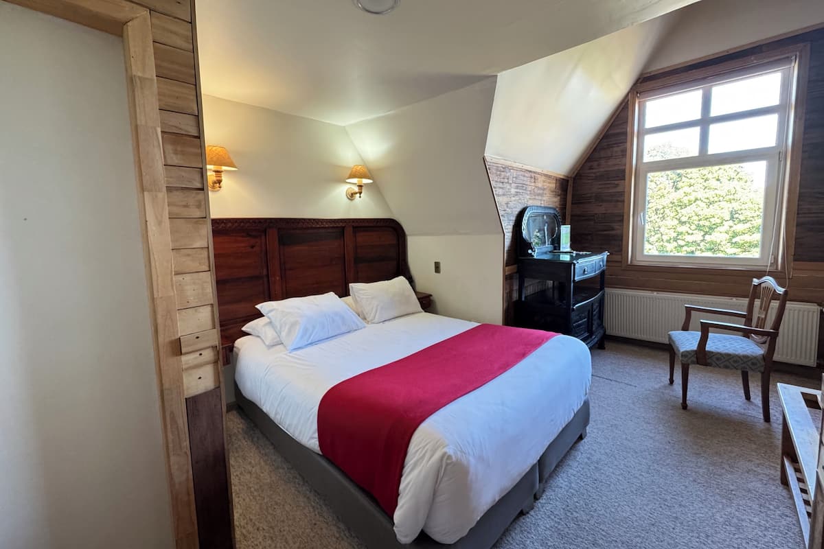 A bedroom with a wooden headboard, a bed with white bedding and a red blanket, a nightstand, a dresser, a chair, and a window with a view of greenery.
