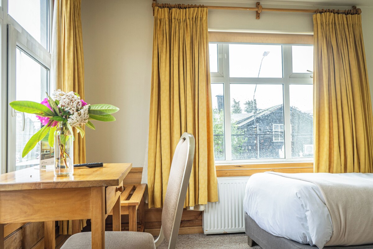 Een kamer met een bed, een bureau met een bloevas, een stoel en een raam met gordijnen.