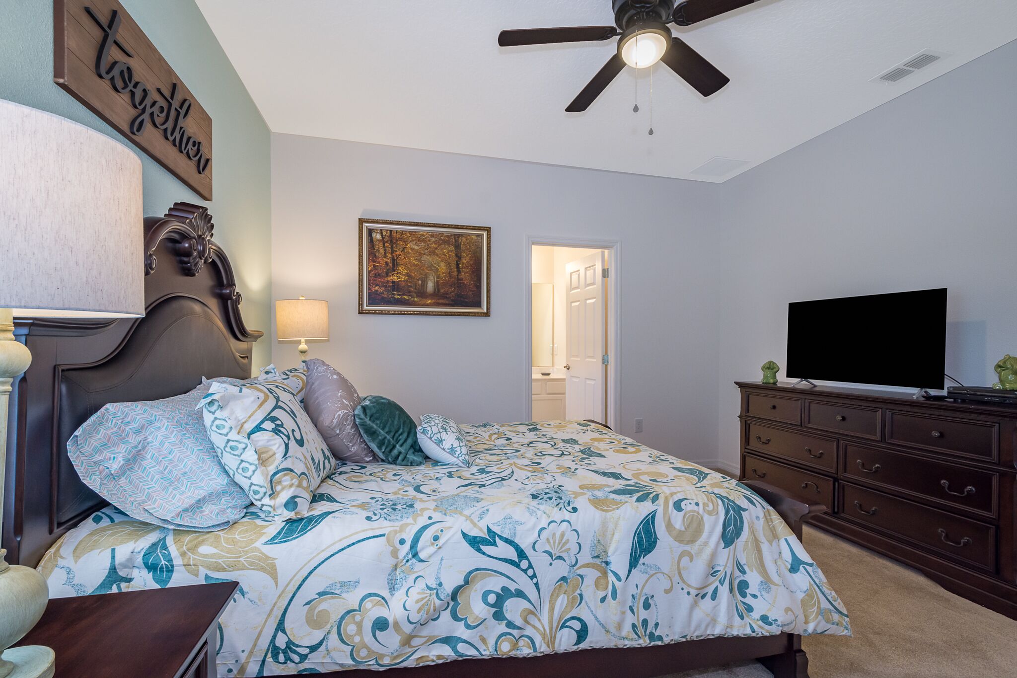 A bedroom with a bed, a ceiling fan, a television on a dresser, and a door leading to another room.