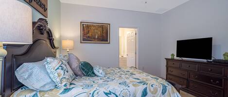 A bedroom with a bed, a ceiling fan, a television on a dresser, and a door leading to another room.