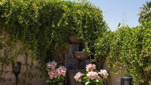 Ein Garten mit Kübeln voller pinkfarbener Blumen, einem Steinbrunnen und einem schwarzen Terratherm.