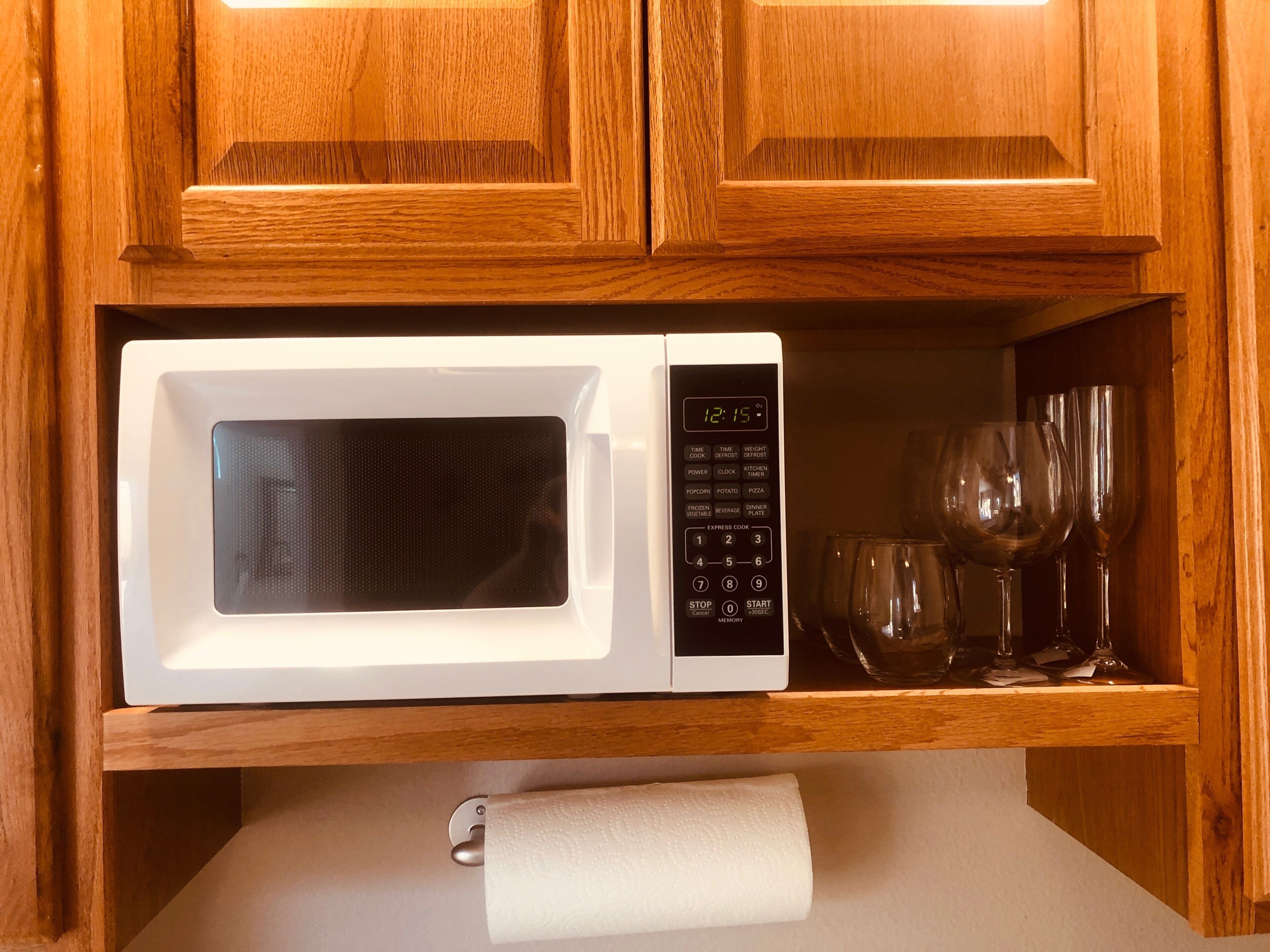 Un four à micro-ondes blanc, doté d’un écran numérique et d’un panneau de commande, est posé sur une étagère en bois, au-dessus d’un porte-rouleau de papier toilette.