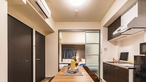 Habitación de hotel compacta con una mesa de comedor de madera, una kitchenette, una cama con cabecera azul y una puerta corrediza de vidrio que da a un balcón.