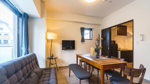 Una sala de estar moderna con un sofá, una mesa de comedor con sillas, un televisor y una cocina visible a través de una puerta abierta.