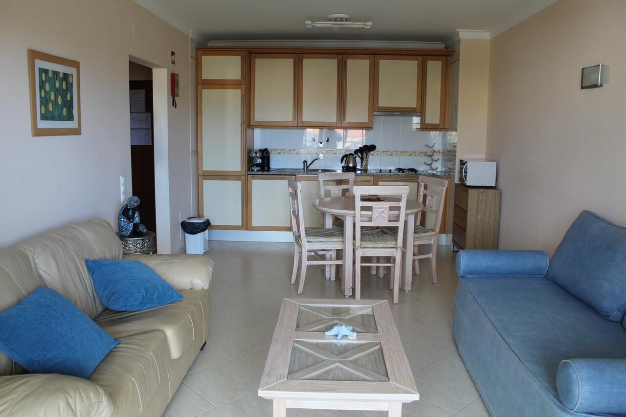 Un salon avec un canapé beige, un canapé bleu et une table basse en verre. On y trouve également un coin repas avec des chaises et une table en bois. La cuisine est visible au fond, avec des meubles en bois et un four à micro-ondes.
