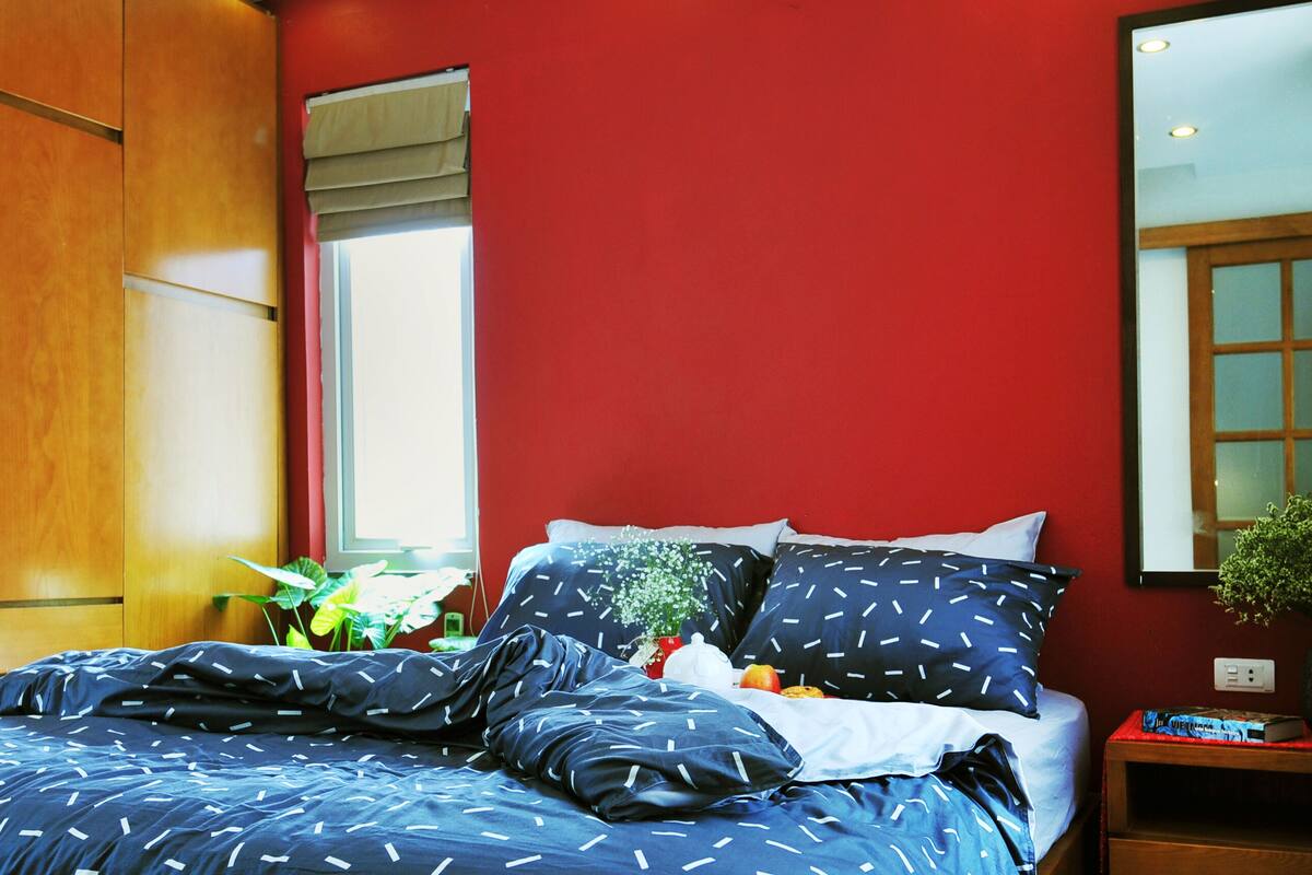 A bedroom with a red wall, a wooden wardrobe, a bed with blue bedding, a window with a view, and a bedside table with a plant.