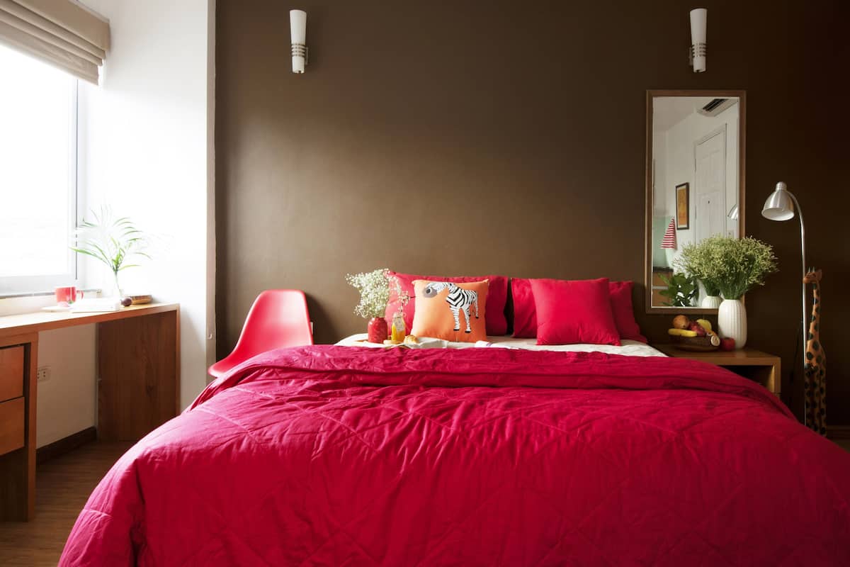 A bedroom with a large bed, a desk, and a chair. There is a window with blinds, a mirror, and a lamp.