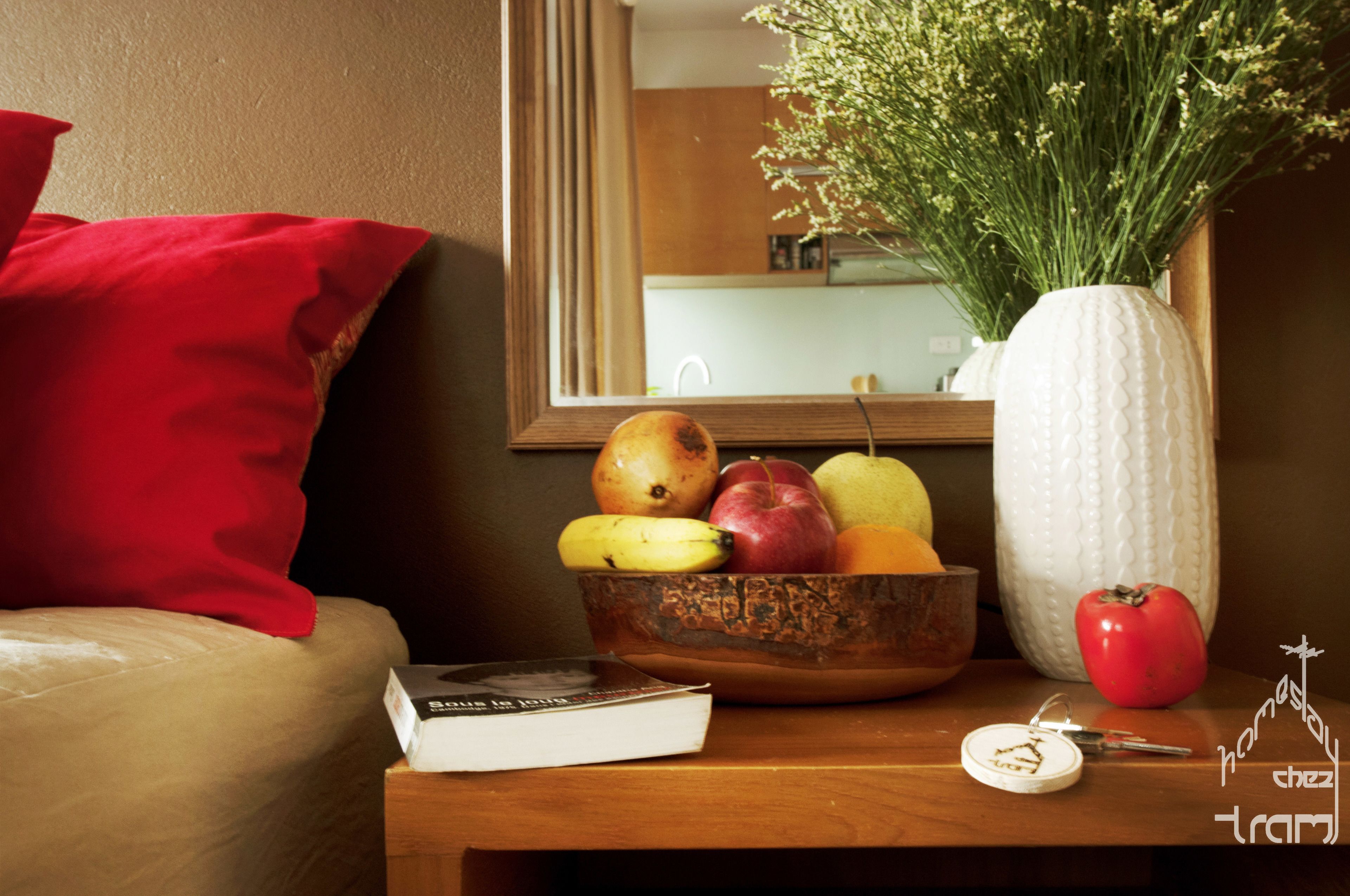 A hotel room with a wooden nightstand, a red pillow, a vase with green plants, a fruit bowl with apples, pears, bananas, and a book.