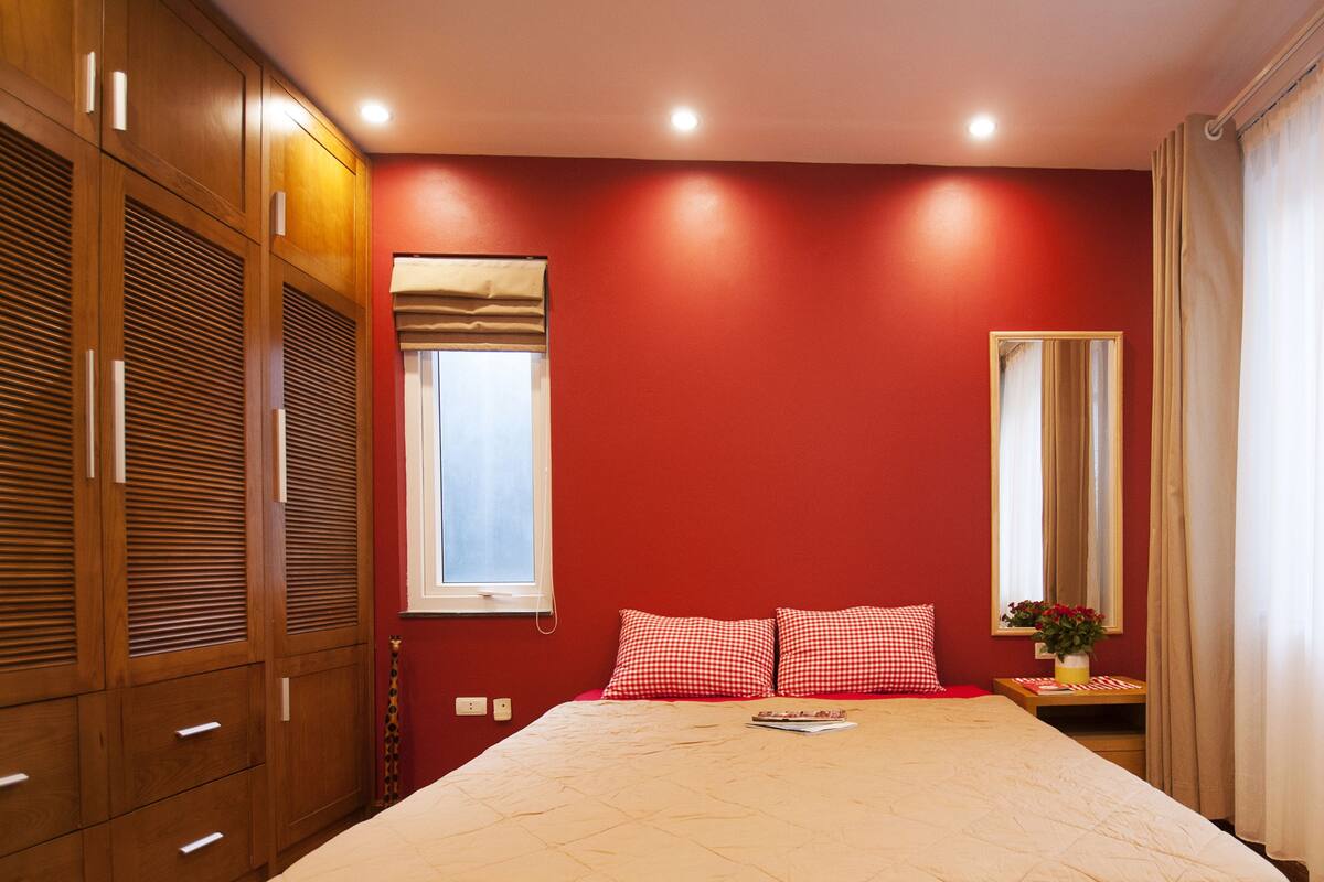 A bedroom with a bed, two checkered pillows, a bedside table with a vase, a mirror, and wooden cabinets.