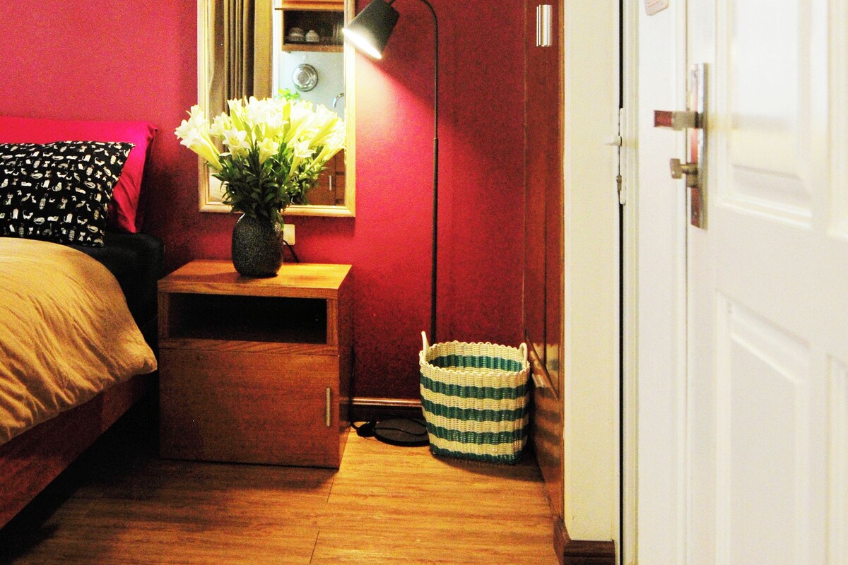 A bedroom with a bed, a nightstand, a lamp, a vase with flowers, and a door.