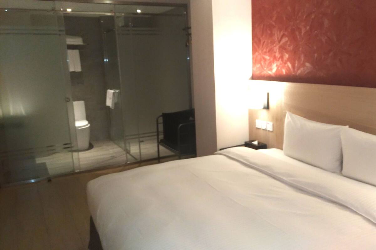 A hotel room with a bed, a nightstand, a wall-mounted lamp, and a glass-enclosed shower area.