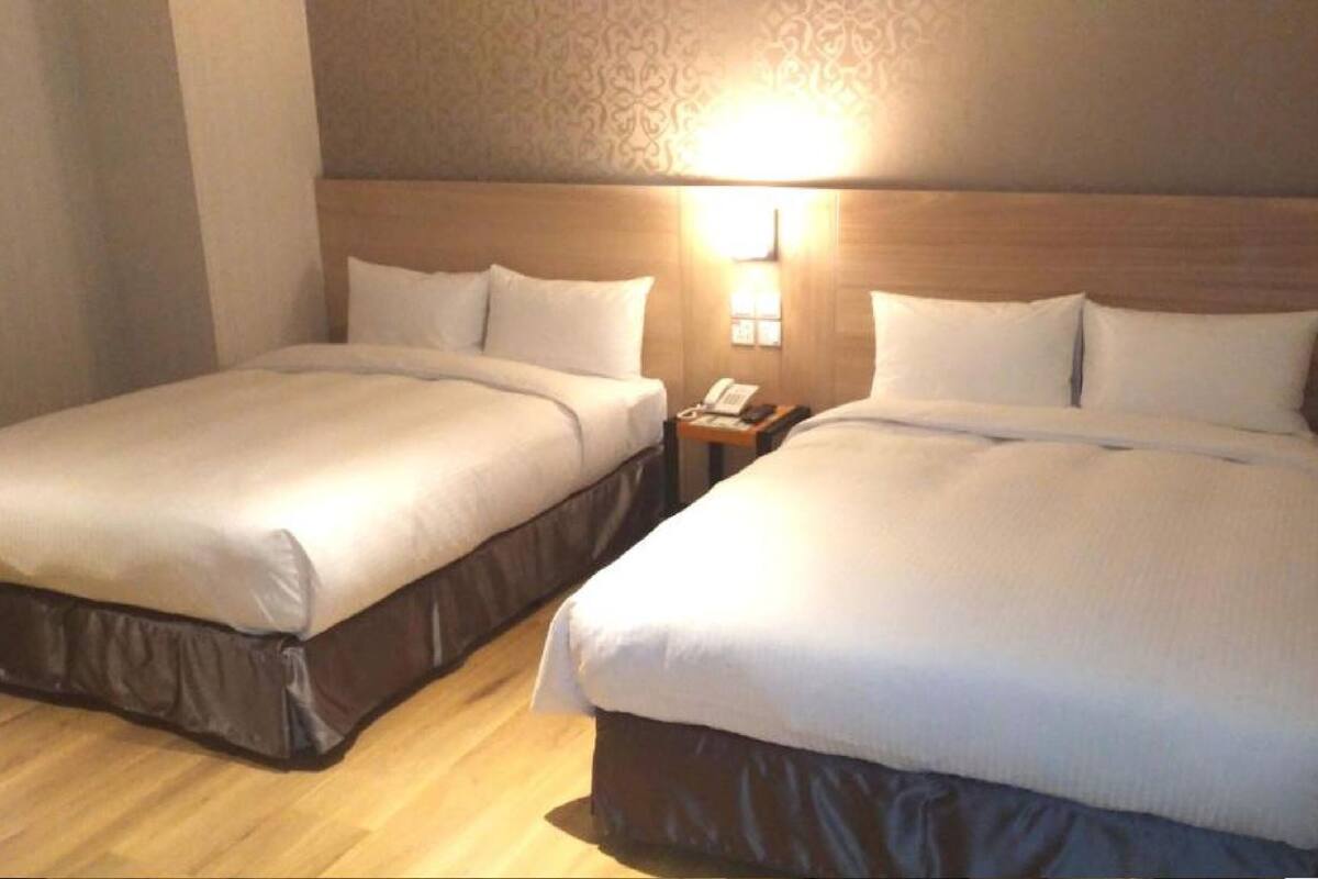A hotel room with two beds, a nightstand with a phone, and a wall-mounted light.