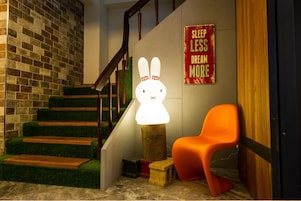 Una escalera con detalles de madera y peldaños cubiertos de moqueta verde, una lámpara blanca y brillante fijada a la pared, una silla naranja y un letrero que dice 'SLEEP LESS DREAM MORE'.