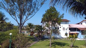 A white building with multiple windows and a red roof, surrounded by greenery and trees. - Apartamento Piccola Porto Bracuhy (Angra dos Reis)