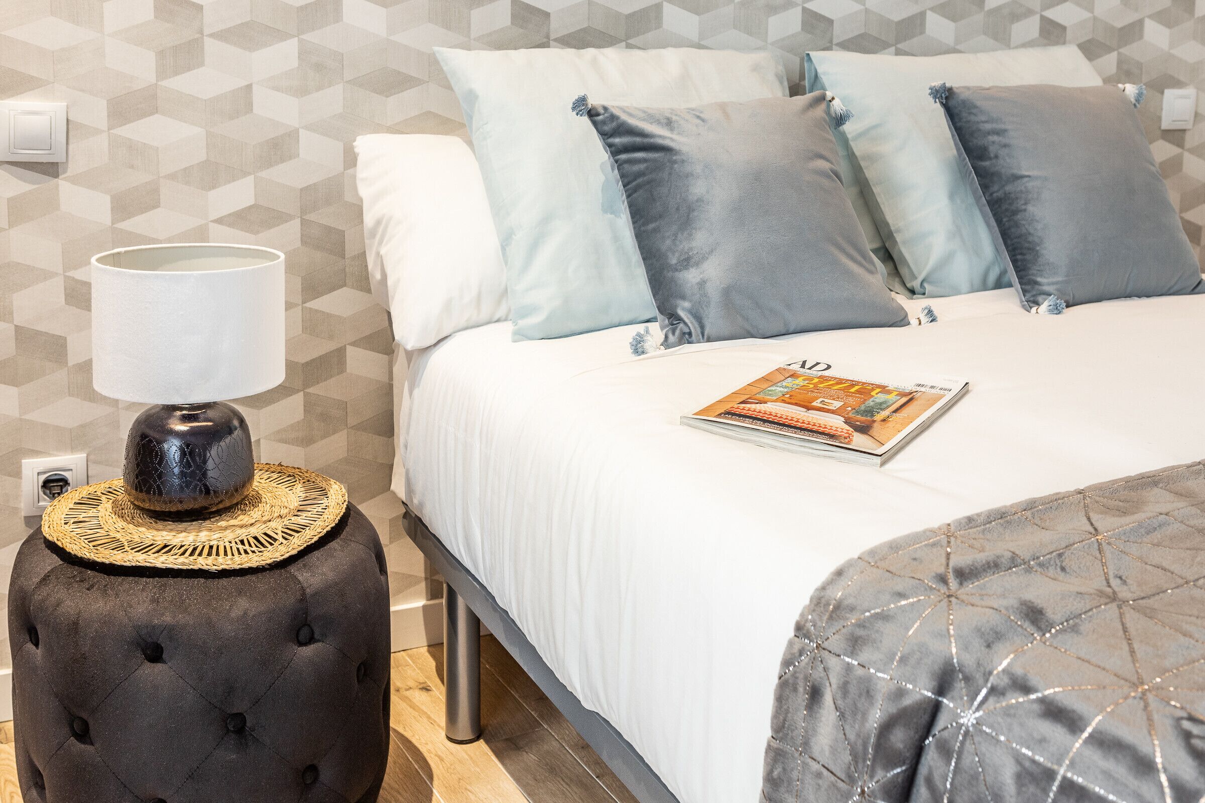 Una cama bien hecha con una revista, una lámpara sobre un taburete y una pared con estampado geométrico.