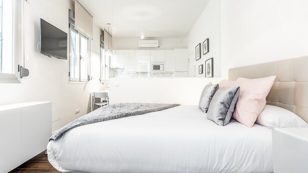 Un dormitorio moderno con una cama grande, un televisor montado en la pared y una cocina visible a través de una puerta abierta.