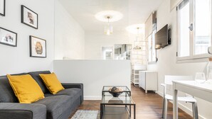 Una sala de estar moderna con un sofá gris, cojines amarillos, una mesa de centro de vidrio y una zona de comedor blanca con un televisor montado en la pared.