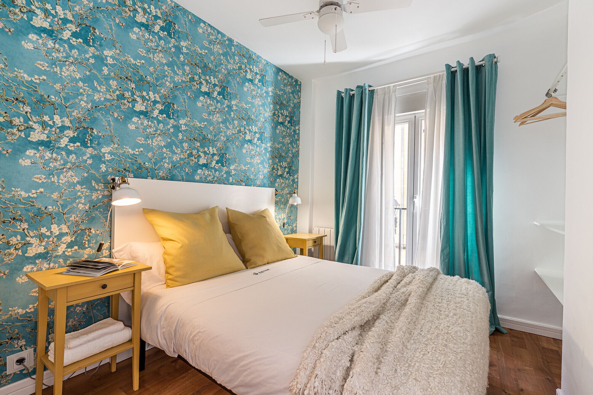 A bedroom with a bed, a bedside table, a lamp, a window with curtains, and a wall with floral wallpaper.