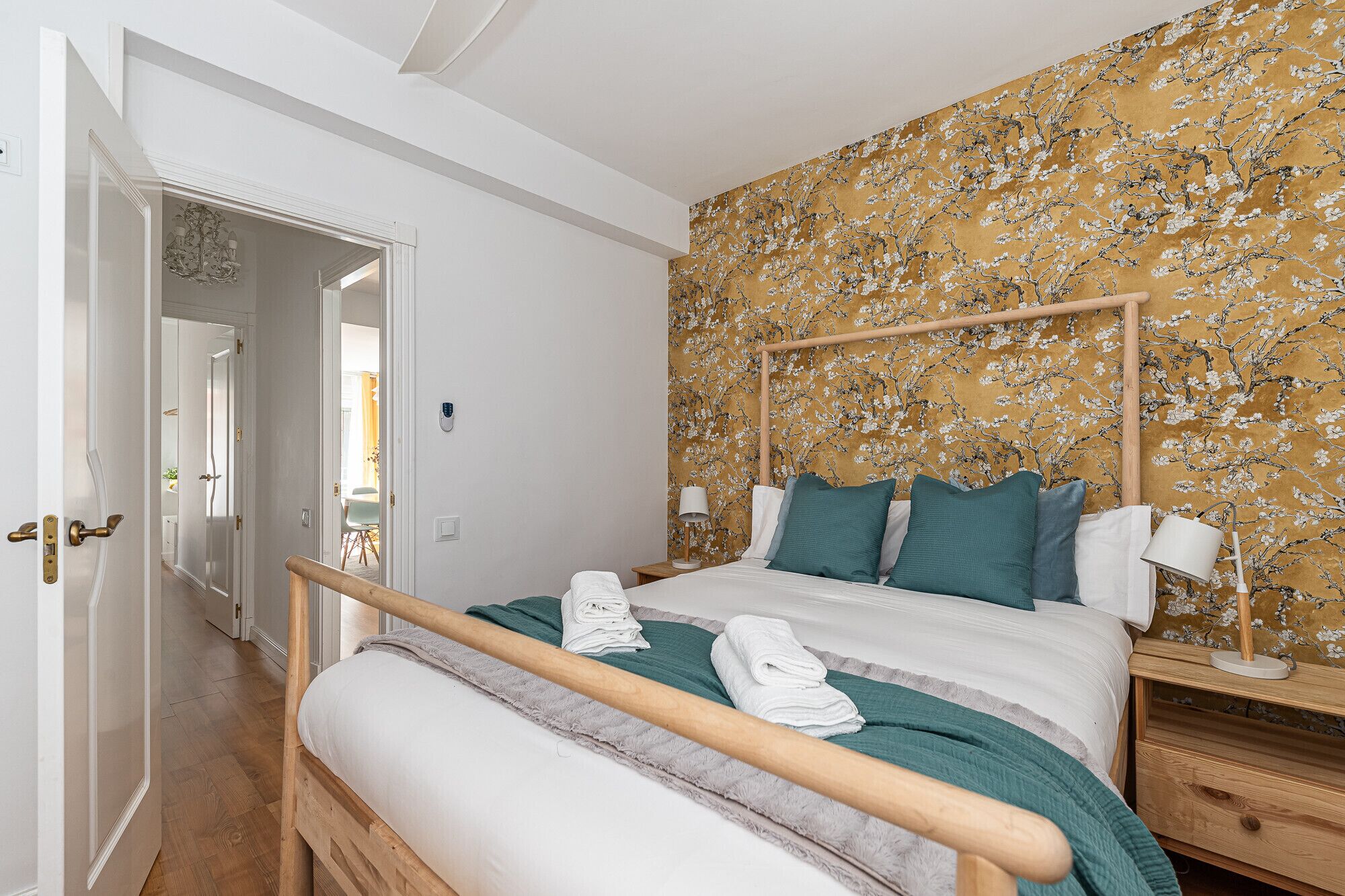 Un dormitorio con estructura de cama de madera, ropa de cama blanca y almohadas turquesa. Hay una mesita de noche de madera con una lámpara y una luz empotrada en la pared. Se aprecia un papel tapiz floral en la pared.