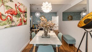 A dining area with a white table, light blue chairs, and a vase with flowers. There is a painting of flamingos on the wall and a mirror on the right. - Waou La Latina II - Alquiler Corta Duración (Madrid)