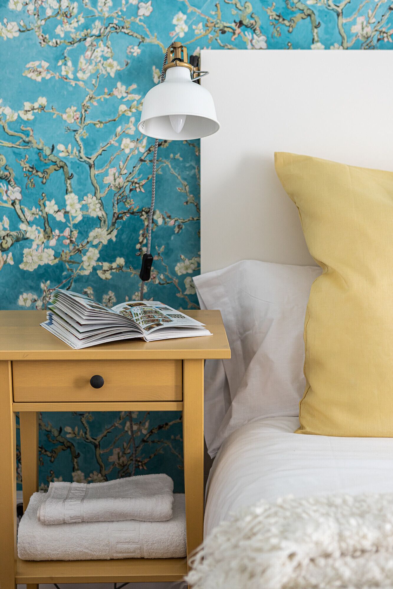 A neatly made bed with a white and yellow pillow, a bedside lamp, and a stack of magazines on a wooden nightstand.