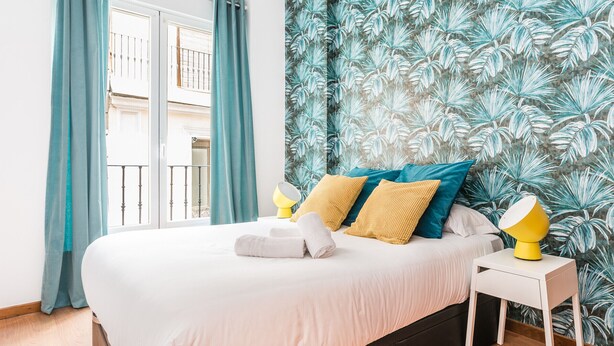 A bedroom with a bed, a nightstand, a window with curtains, and a wall with a leaf pattern. - Waou Gran Via I - Alquiler Corta Duración (Madrid)