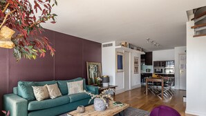 Un salon moderne avec un canapé de couleur turquoise, une table basse en bois et un pouf violet. On aperçoit une cuisine à l’arrière-plan, équipée d’appareils électroménagers en acier inoxydable.