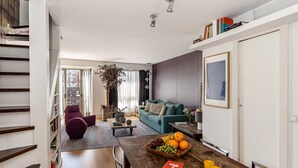 Un salon moderne avec un canapé vert, une table basse en bois sur laquelle sont disposés des fruits et des boissons, un fauteuil violet et un escalier agrémenté d’étagères à livres.