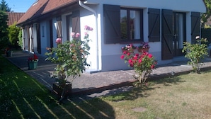 A white house with a brown roof, large windows with shutters, and potted plants with pink flowers.