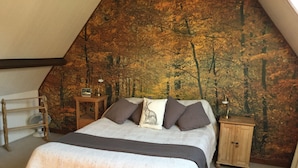 A bedroom with a sloped ceiling, a bed with a white and brown blanket, multiple pillows, a small wooden nightstand with a lamp, and a wall mural of a forest.