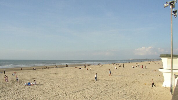 A wide sandy beach with people scattered across it, a lifeguard tower in the background, and the ocean in the distance.
