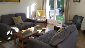 A living room with a sofa, armchair, and a small table. There is a door leading to a balcony with a view of the street.