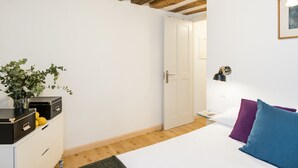 Quarto com teto de madeira, guarda-roupa branco, cama com almofadas azuis e roxas e uma pequena mesa com abajur.