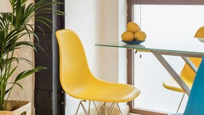 Um quarto com uma mesa de vidro, cadeiras amarelas, um vaso de planta e uma janela grande.