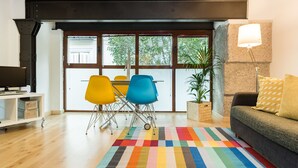 Uma sala de estar moderna com teto de madeira, tapete listrado e colorido, sofá com abajur, e um espaço de jantar com cadeiras e mesa.