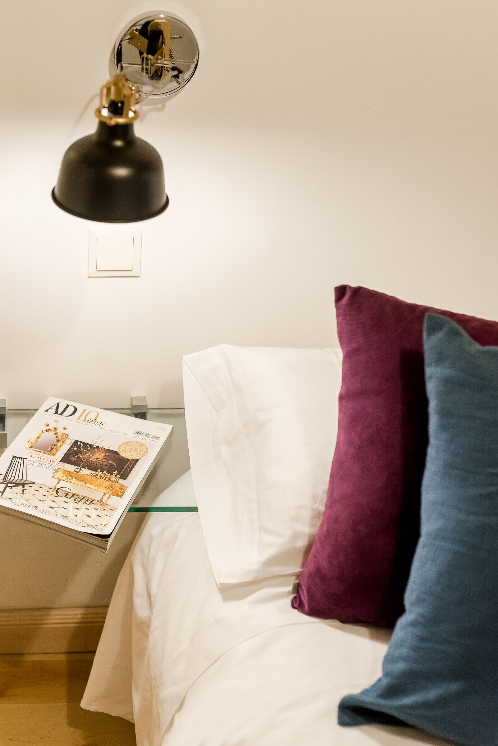 Un lit bien fait, agrémenté d’oreillers blancs et colorés, un magazine posé à proximité et une lampe de bureau noire.