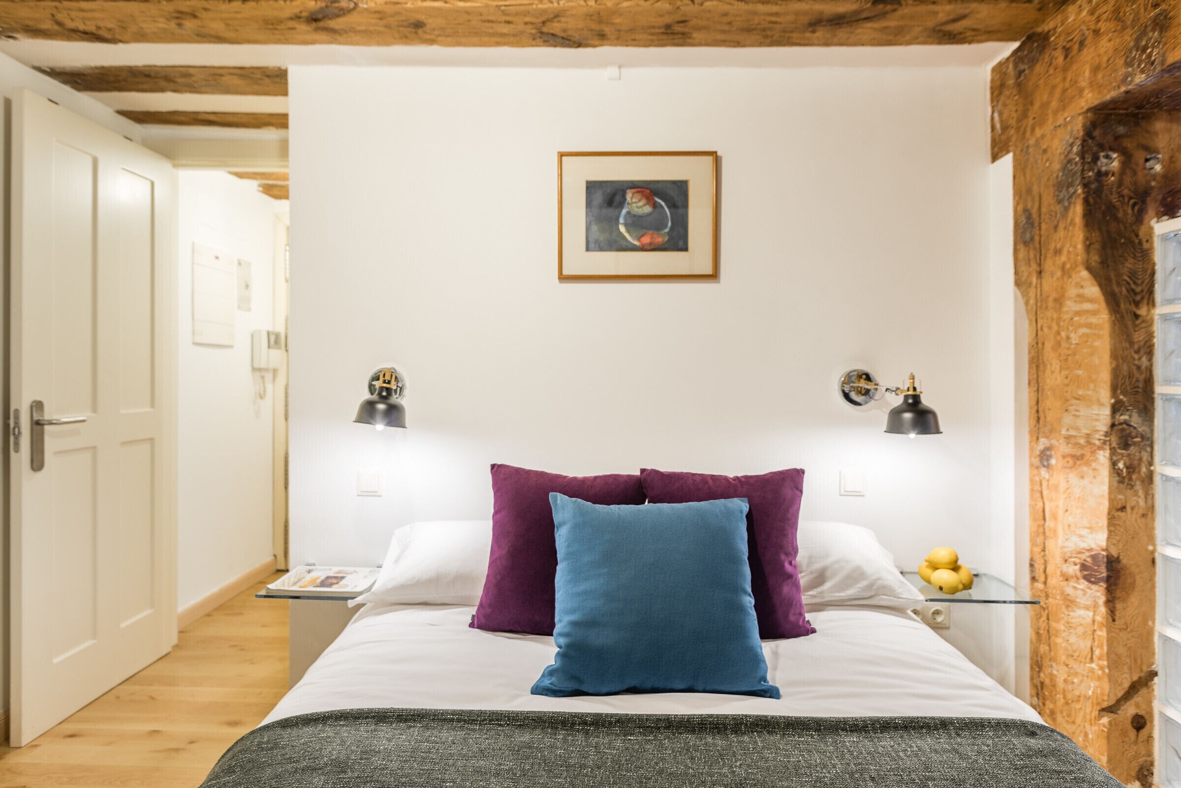 Un dormitorio con una cama, almohadas, una mesita de noche, una lámpara y un cuadro colgado en la pared.