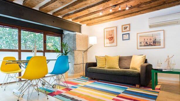 Uma sala de estar moderna com teto de madeira, tapete listrado e colorido, sofá cinza com almofadas amarelas, e um espaço de jantar com cadeiras amarelas.