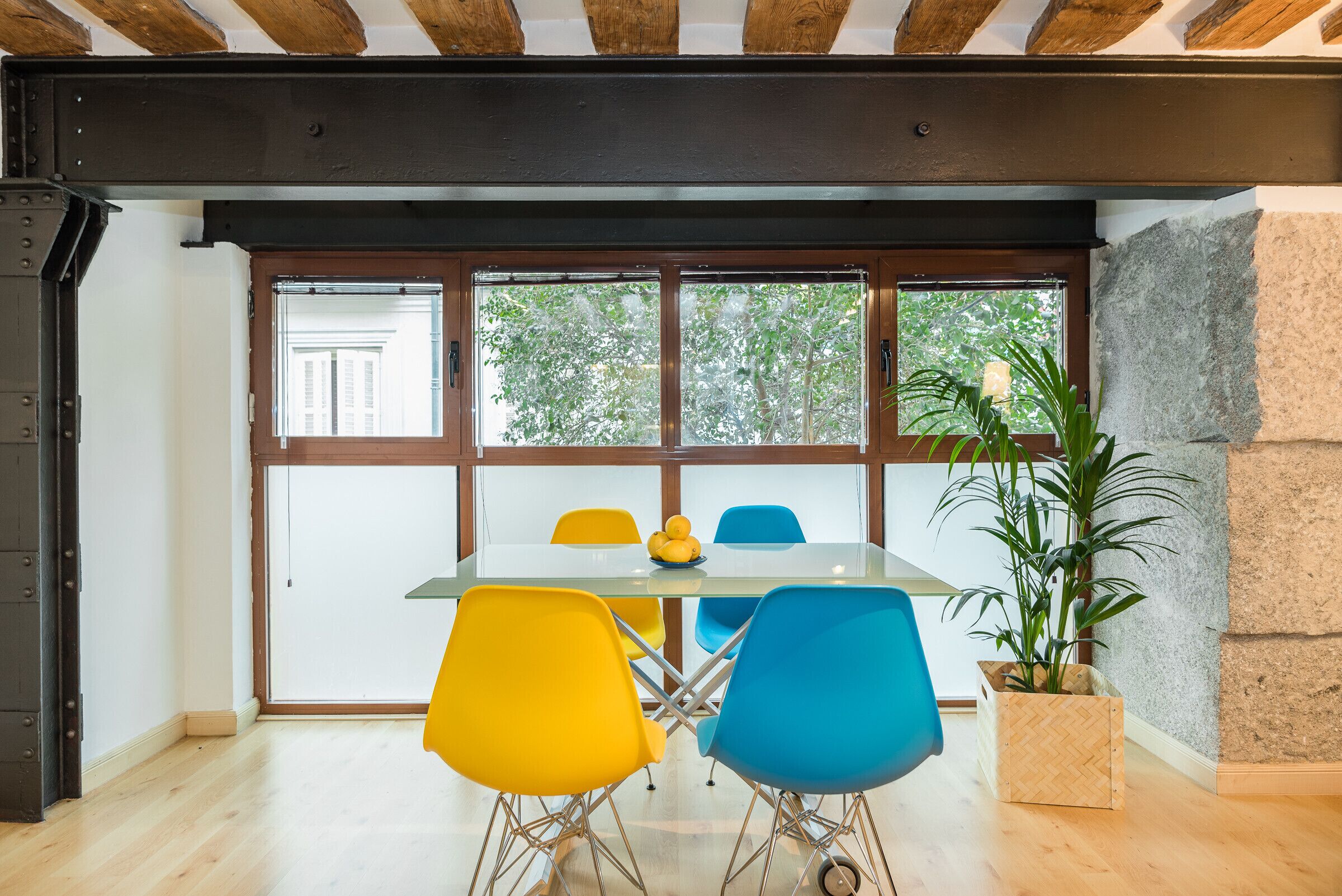 Un coin repas avec une table en verre, des chaises jaunes et bleues, une plante en pot et une vue dégagée grâce à de grandes fenêtres.