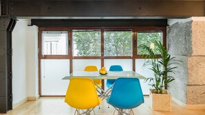 Uma sala de jantar com mesa de vidro, cadeiras amarelas e azuis, um vaso de plantas e vista através de grandes janelas.