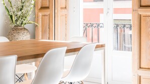 Comedor con una mesa de madera y sillas blancas, un florero con flores y vistas a un balcón con plantas.