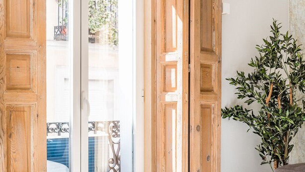 Una habitación con puerta de madera, un sofá gris, una mesa redonda pequeña y una planta en maceta.