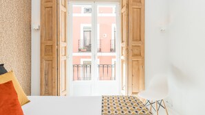 Una habitación con una cama, una silla blanca y un tapete estampado. Se observa una puerta de madera con paneles de vidrio que da a un balcón exterior.