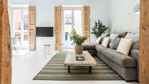 Una sala de estar moderna con un sofá gris, una mesa de centro de madera con un libro, un tapete verde estampado y un ventanal con contraventanas de madera.