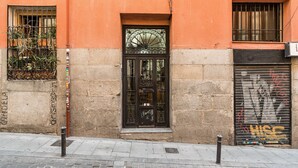 Fachada de edificio con una puerta negra grande, una ventana con barrotes metálicos y un toldo cerrado con grafitis.