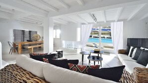 A spacious living room with a large window overlooking a pool and a scenic view. - Villa Blanca Uno (Mykonos)