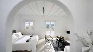 A white room with a bed, a chair, a table, and a view of the sea through a window. - Villa Blanca Uno (Mykonos)