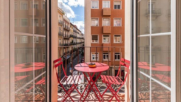 Een balkon met rode stoelen en een kleine tafel, uitkijkend op een straat met gebouwen en een heldere lucht.