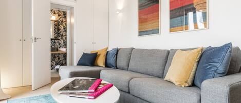 Una sala de estar moderna con un sofá seccional gris, una mesa de centro blanca con libros, y un cuadro abstracto y colorido en la pared.